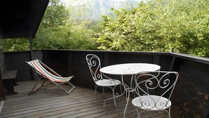 Een houten terras met een witte ronde tafel en metalen stoelen, met uitzicht op een bebost gebied.
