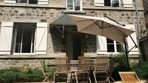 A stone building with multiple windows, a covered patio with wooden furniture, and a lounge chair. - Villa 'L'autre Vie Là' with Mountain Views, Private Terrace and Wi-Fi (La Bourboule)