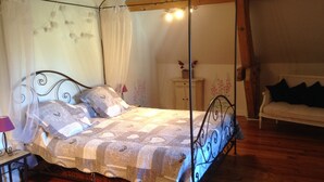 A four-poster bed with a canopy, a bedside table with a lamp, a wooden floor, and a white sofa in the background. - Villa 'L'autre Vie Là' with Mountain Views, Private Terrace and Wi-Fi (La Bourboule)