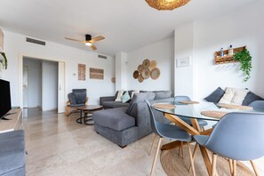 A modern living room with a glass dining table, grey sofas, and a ceiling fan.