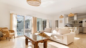 Un vaste espace de vie comprenant une table basse en verre, un canapé blanc, des fauteuils en osier et, au fond, une cuisine.