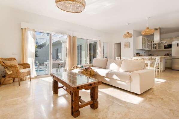 A spacious living area with a glass-top coffee table, a white sofa, wicker chairs, and a kitchen in the background.