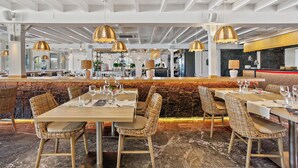 Un restaurant avec de hauts plafonds, des suspensions décoratives et des chaises en osier. Les tables sont dressées avec des nappes blanches et des verres à vin.