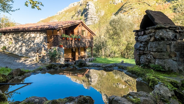 Una cabaña rústica de montaña con chimenea de piedra, balcón de madera con plantas en macetas, un pozo de piedra y un reflejo claro en un pequeño estanque.