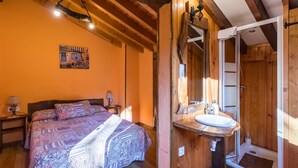 Un dormitorio con cama, cabecera de madera, mesita de noche, cuadro en la pared y un baño de madera con lavamanos, espejo y ducha.