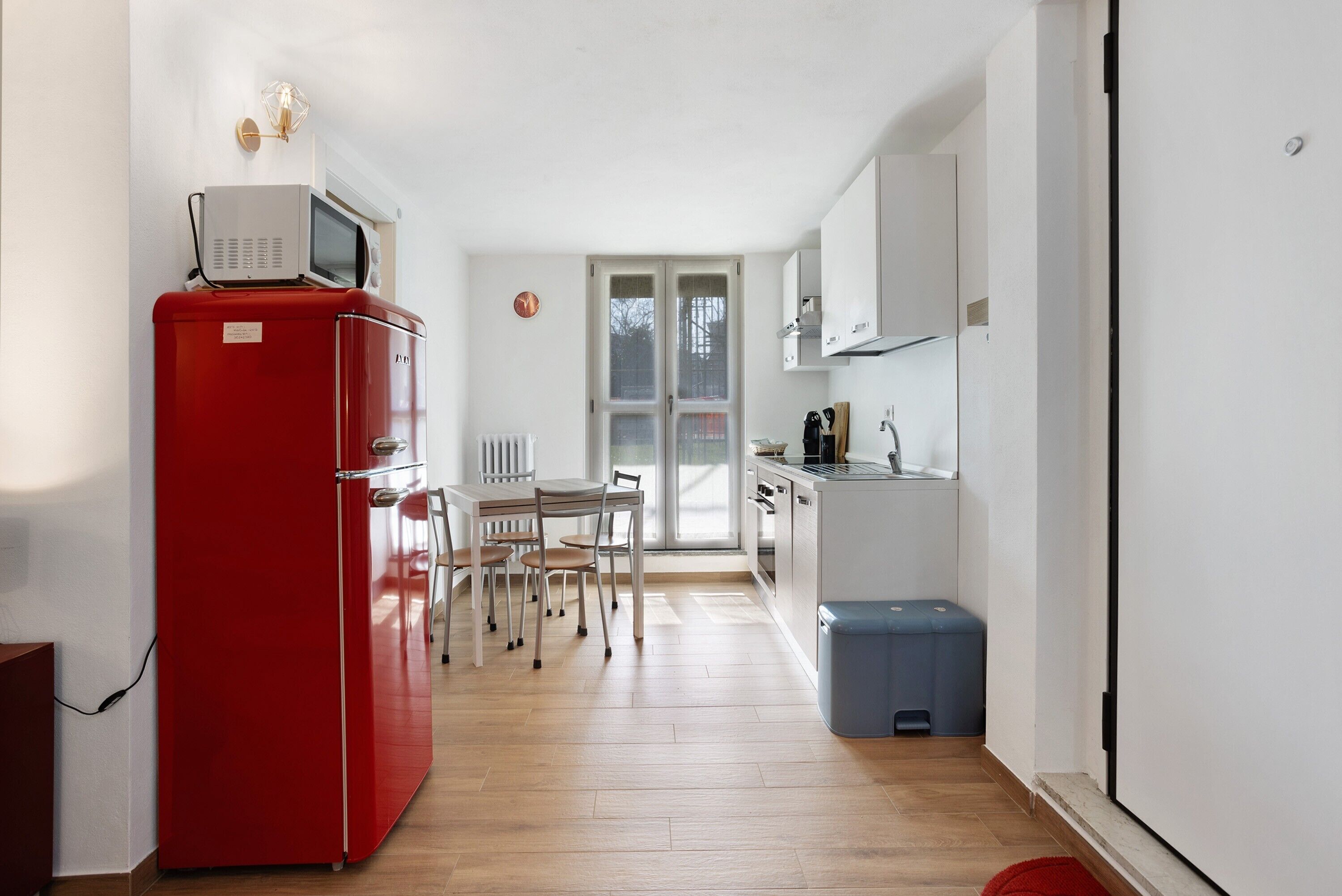 Une cuisine de taille réduite avec un réfrigérateur rouge, une table à manger avec des chaises et une poubelle bleue.