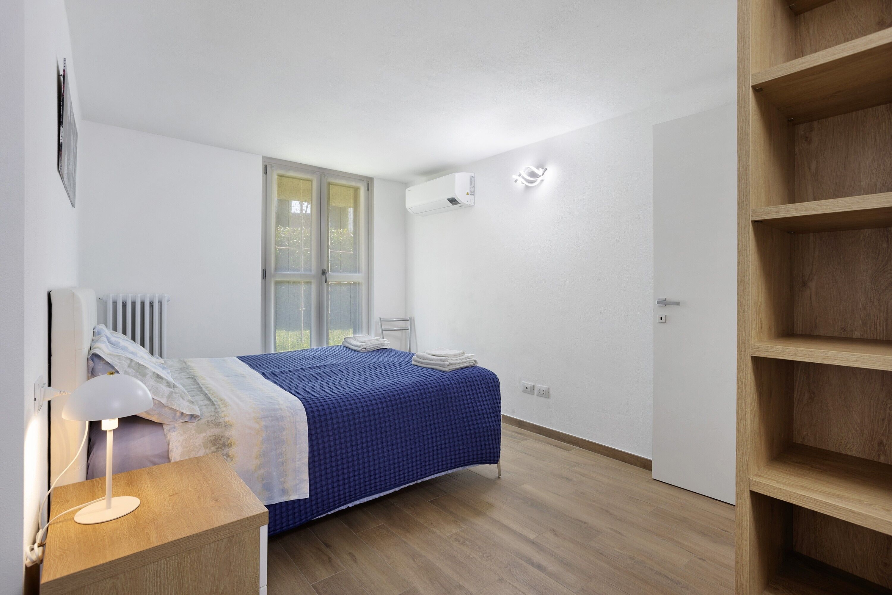 Un dormitorio con cama, mesita de noche, lámpara, radiador, ventana y estanterías empotradas.
