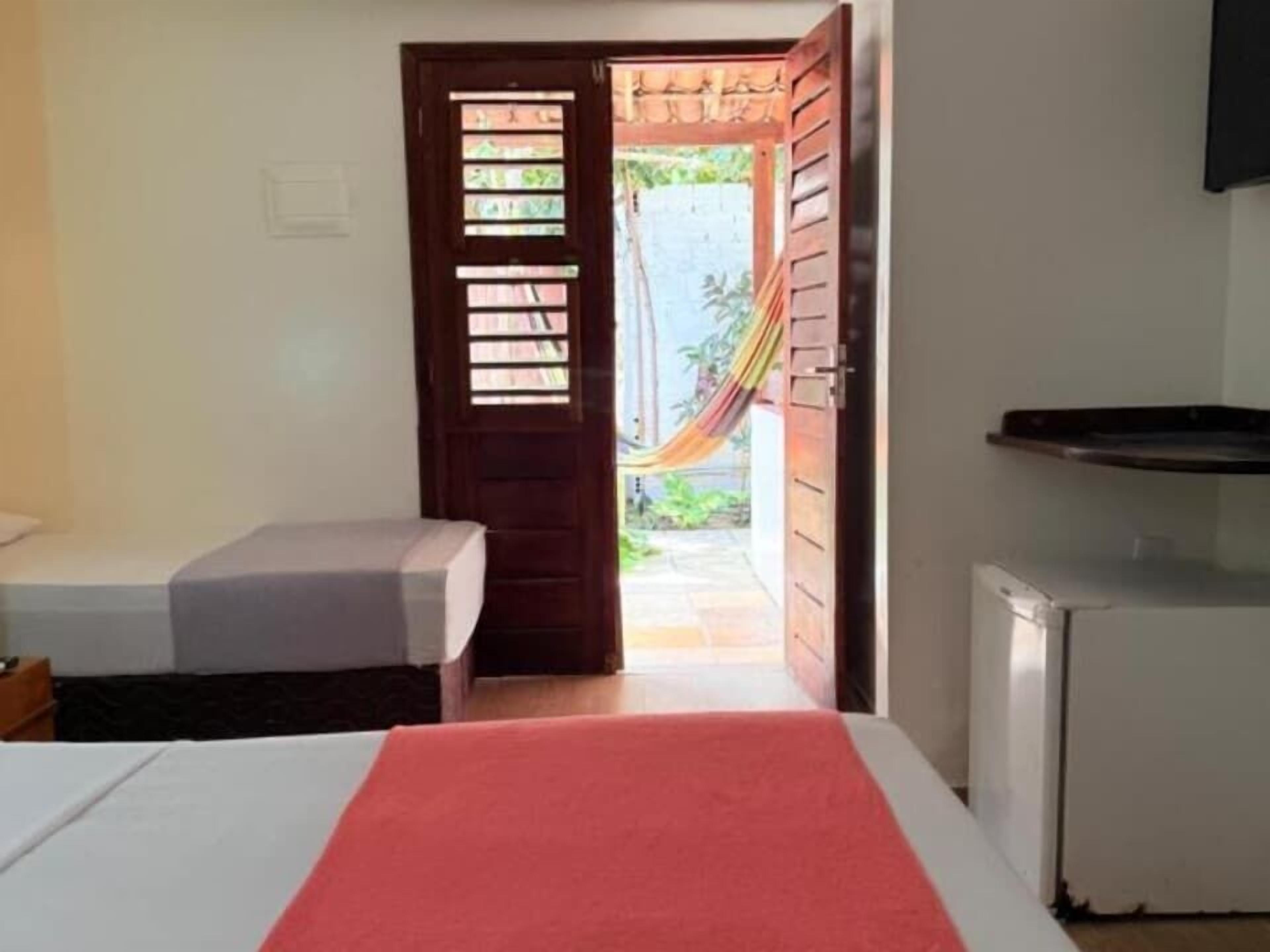 A room with a bed, a small table, a refrigerator, and a door leading to an outdoor area with a hammock.