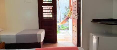A room with a bed, a small table, a refrigerator, and a door leading to an outdoor area with a hammock.