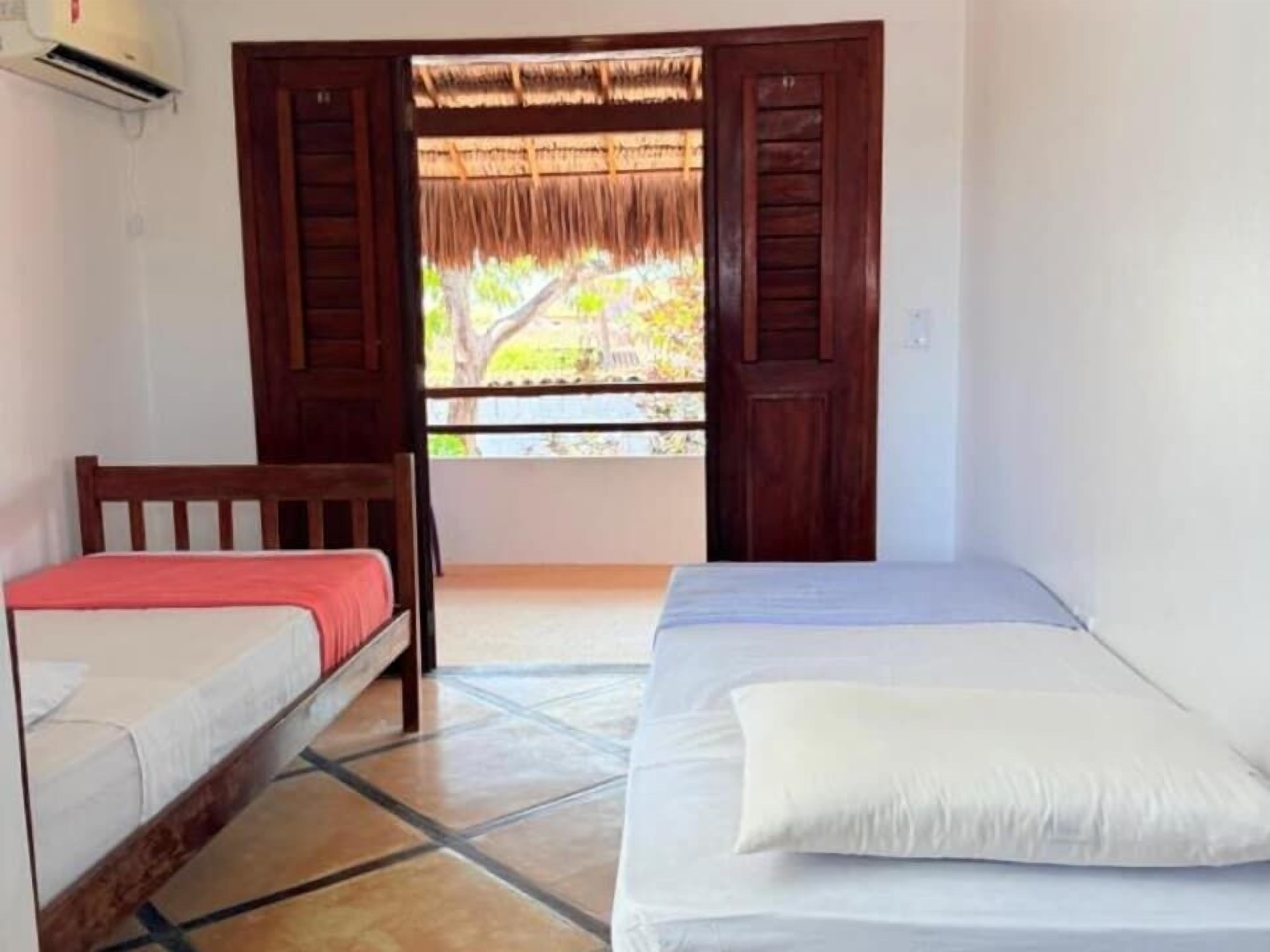 A room with two beds, a window with a view of greenery, and a door with a thatched roof.