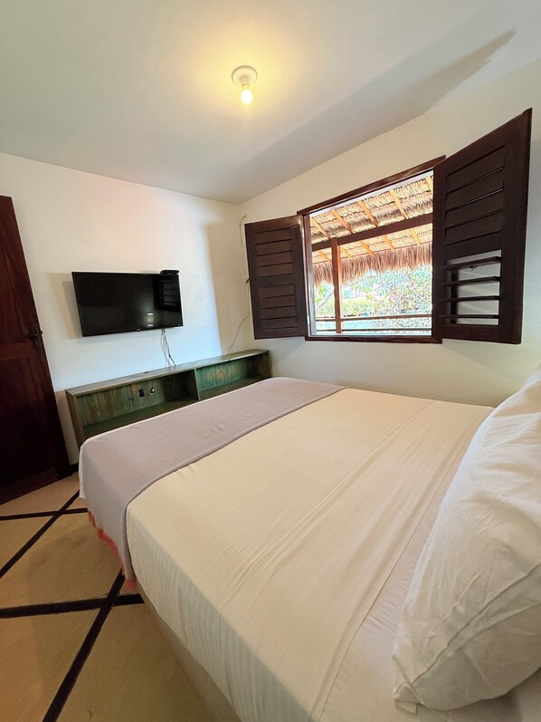 A bedroom with a bed, a TV mounted on the wall, a wooden dresser, and a window with shutters.