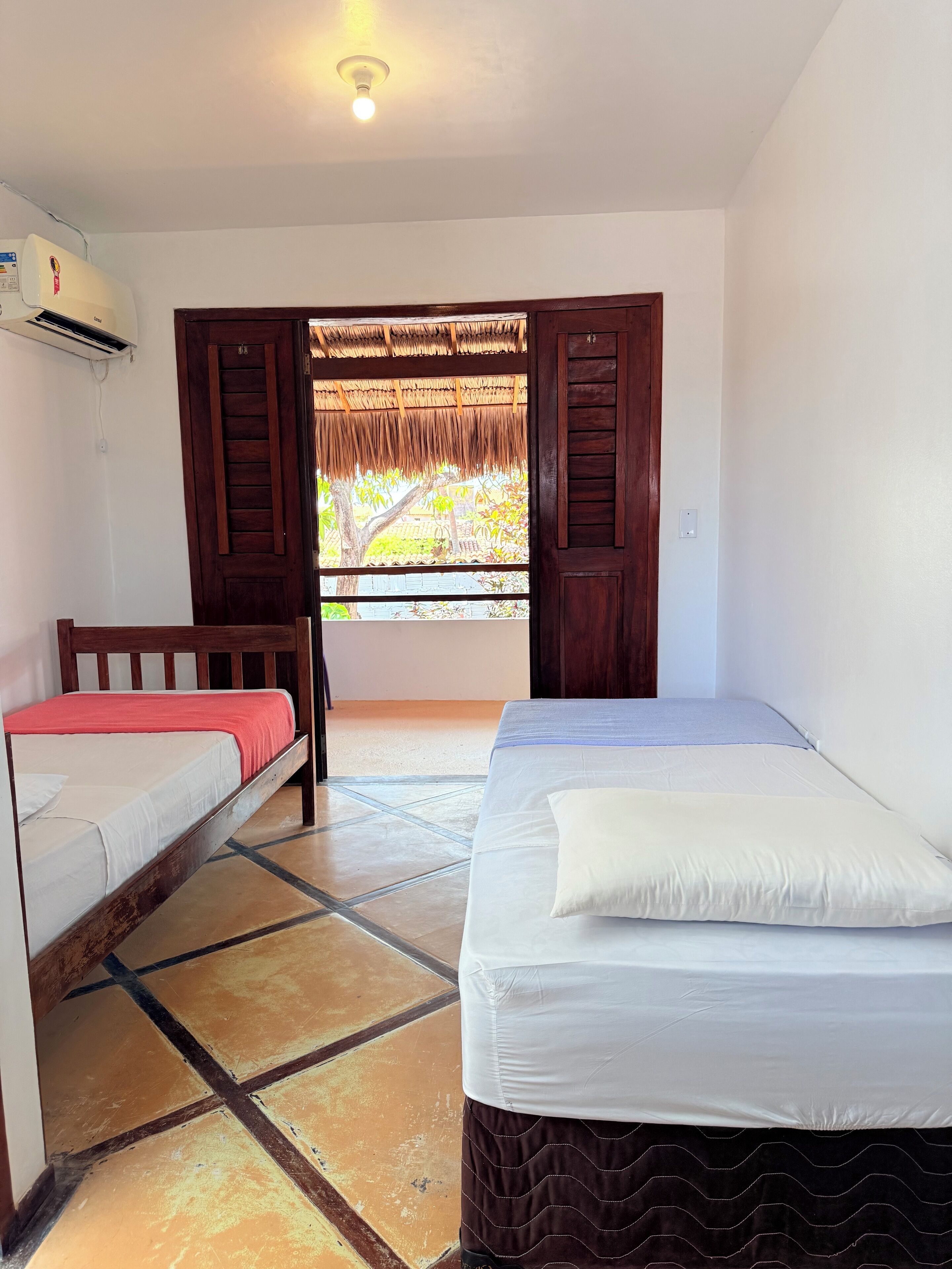 Une chambre avec deux lits, une porte en bois, une fenêtre surmontée d’un toit de chaume et un appareil de climatisation.
