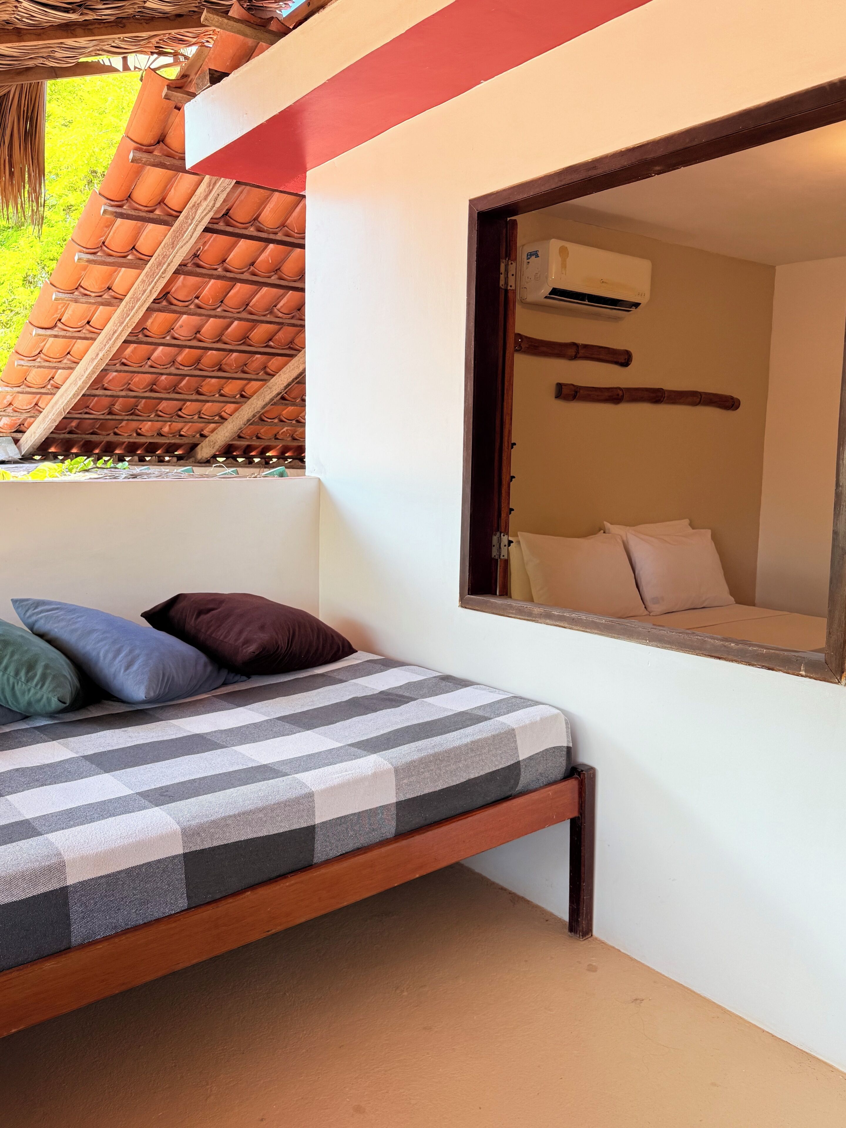 Una cama con colchón a cuadros, almohadas y estructura de madera. Se ve una ventana que da a una habitación con cama y aire acondicionado.