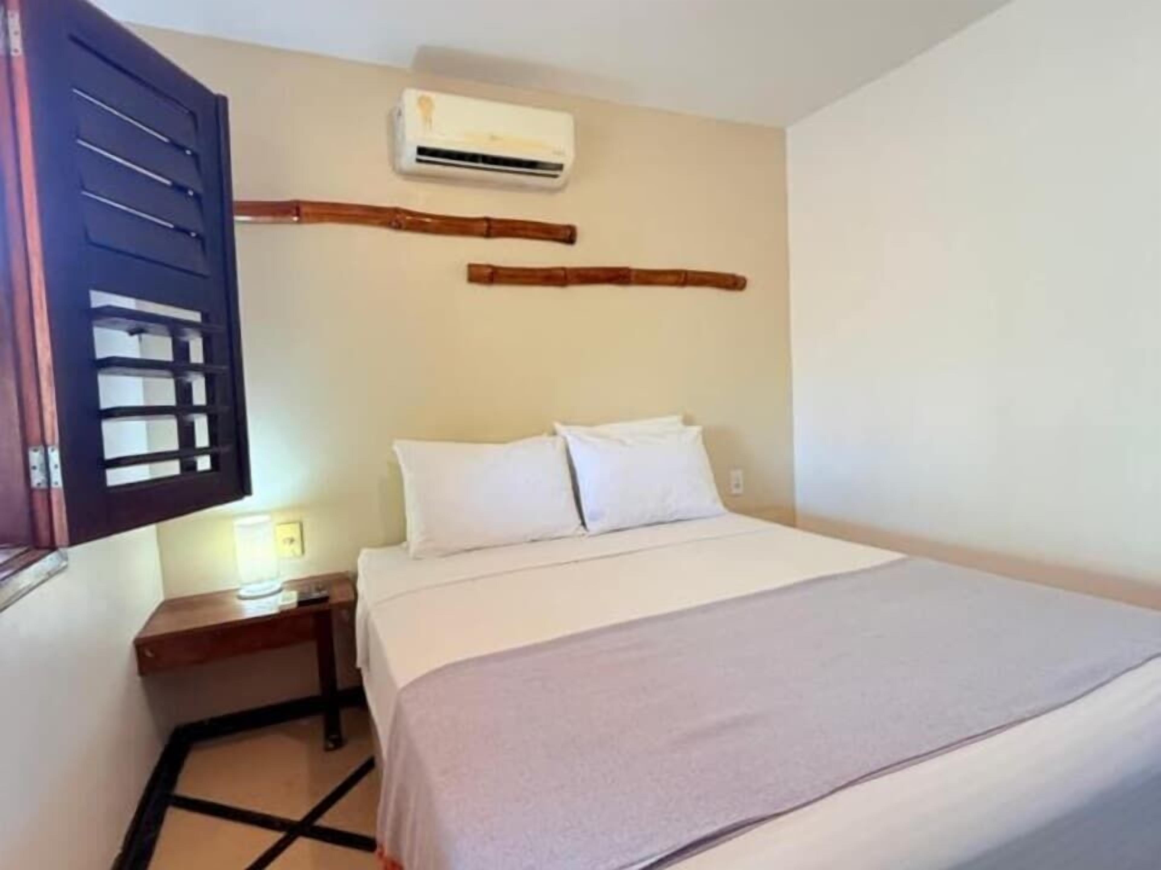 Une chambre d’hôtel comprenant un lit, une table de chevet, un appareil de climatisation et une fenêtre avec un store en bois.