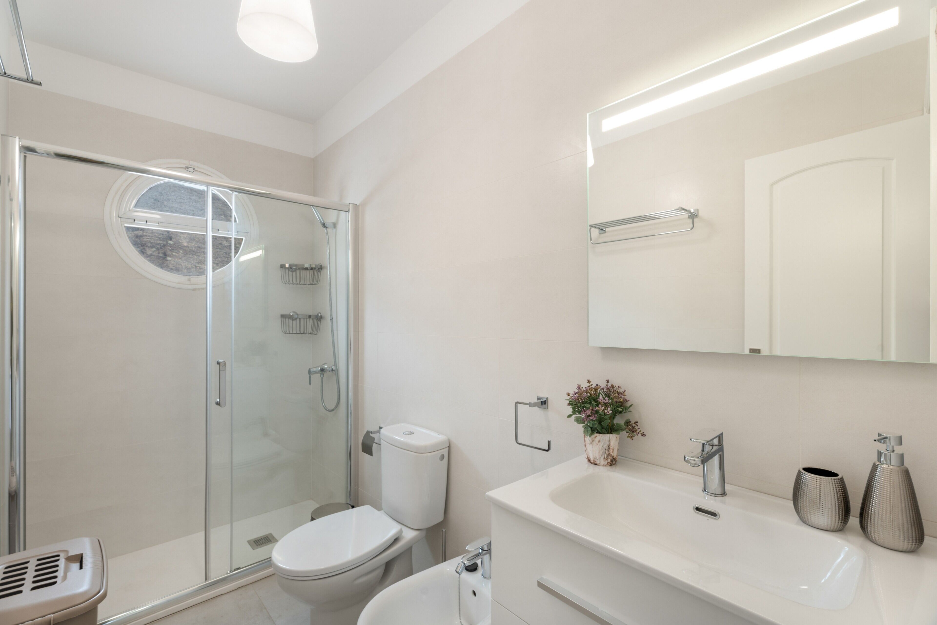 Ein modernes Badezimmer mit Duschkabine aus Glas, Toilette, Waschbecken mit Spiegel darüber und einer kleinen Topfpflanze auf der Ablagefläche.