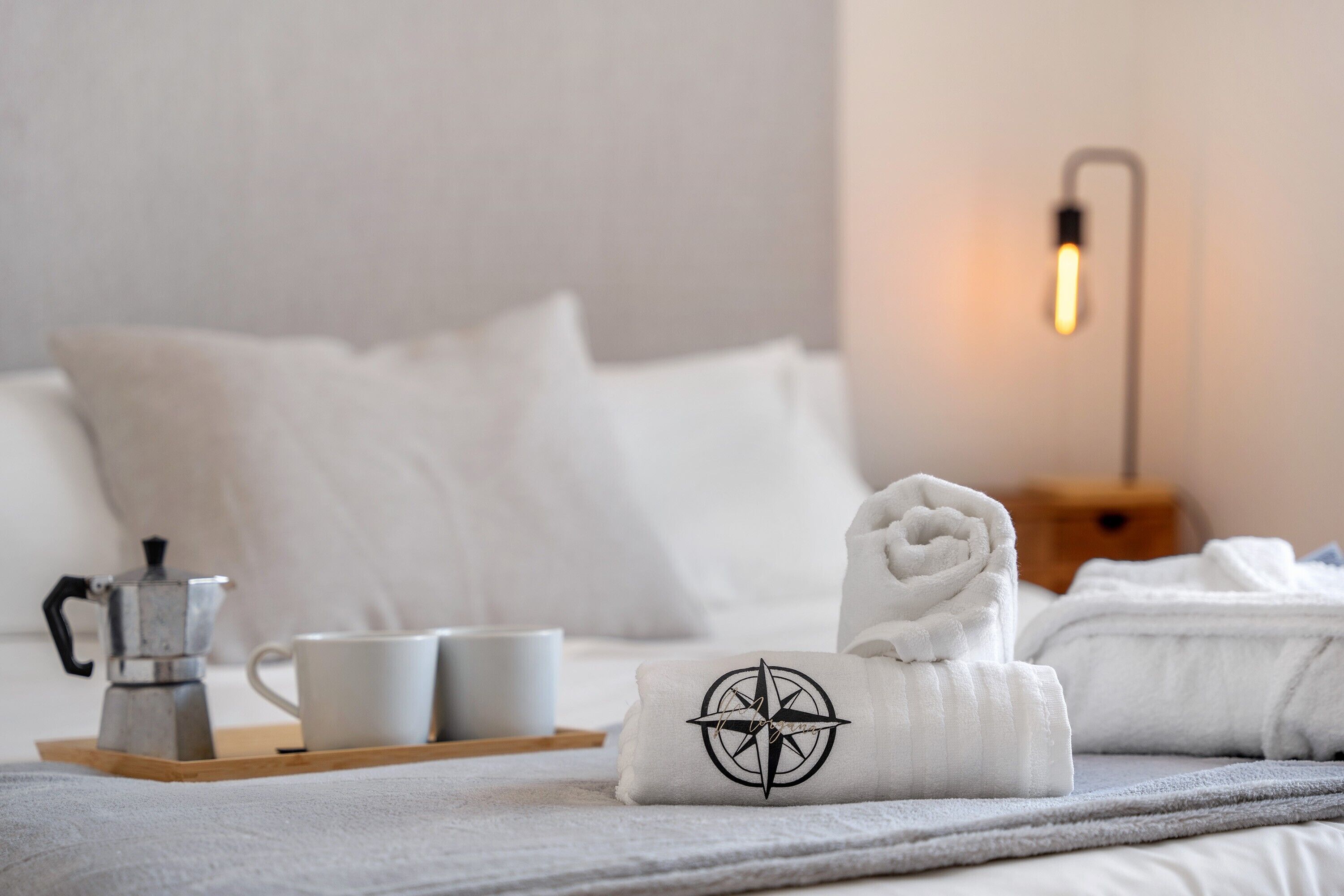 Un lit bien fait, avec du linge de lit blanc, une serviette enroulée ornée du logo d’une boussole, une machine à café et deux tasses blanches sur un plateau.