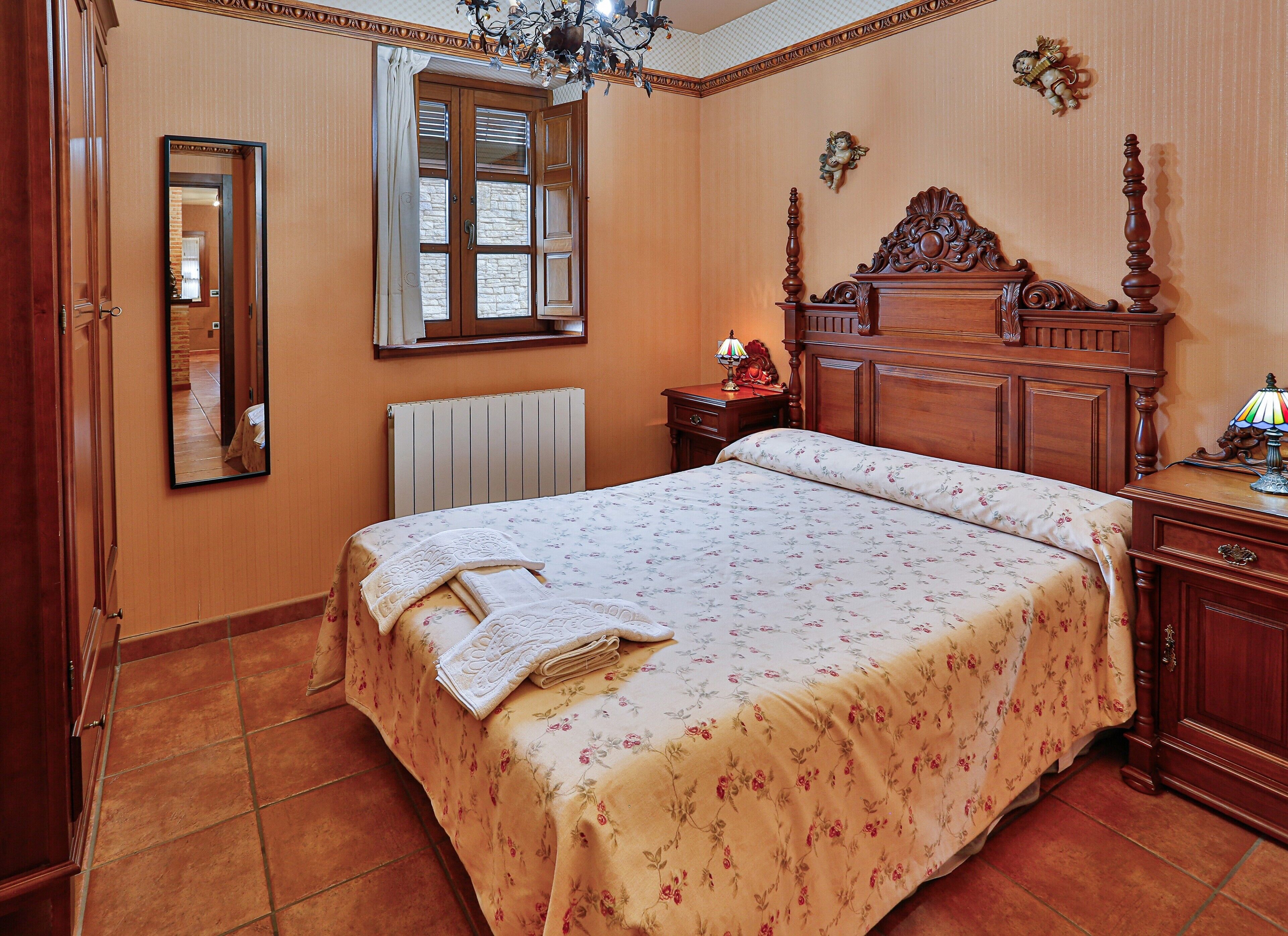 Quarto com uma cama grande, penteadeira, mesa de cabeceira, espelho, janela com cortinas e radiador.