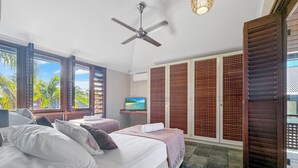 A bedroom with a bed, two bedside tables, a ceiling fan, a TV, and a view of palm trees through large windows. - Wongalinga Beachfront Apt 5 (Wongaling Beach)
