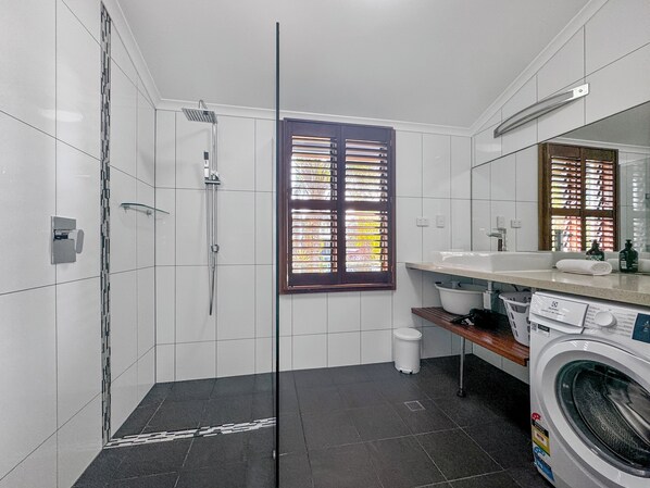 A modern bathroom with a glass shower enclosure, a built-in sink, a washing machine, and a window with blinds.