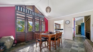 A dining area with a wooden table and chairs, a large window with shutters, and a kitchen in the background. - Wongalinga Beachfront Apt 5 (Wongaling Beach)