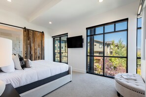 Ein modernes Schlafzimmer mit einem großen Bett, einem Flachbildfernseher und Blick auf eine Wohngegend durch große Fenster.