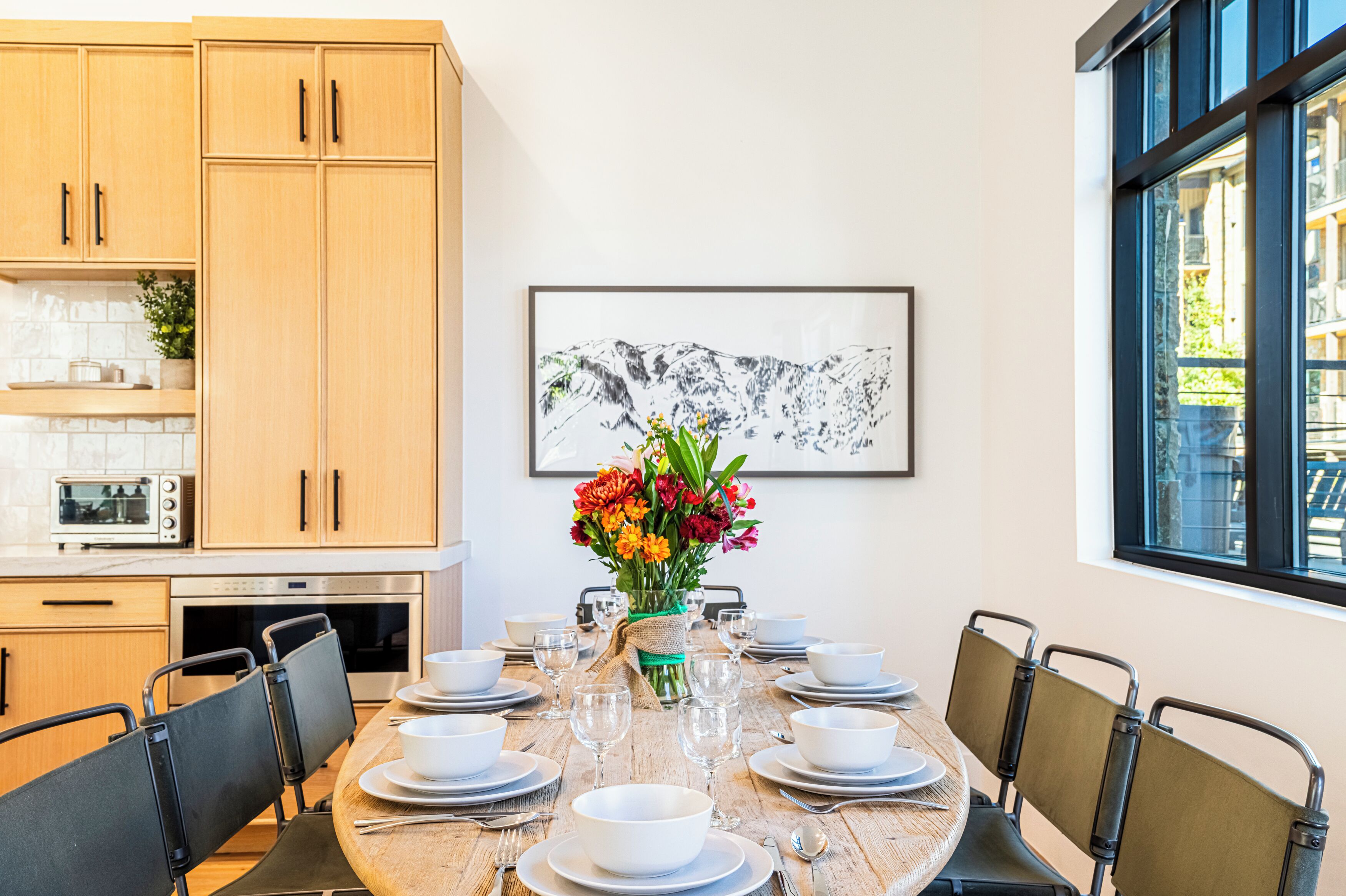 A dining area with a round table set for six, surrounded by chairs. A vase with flowers is placed in the center.