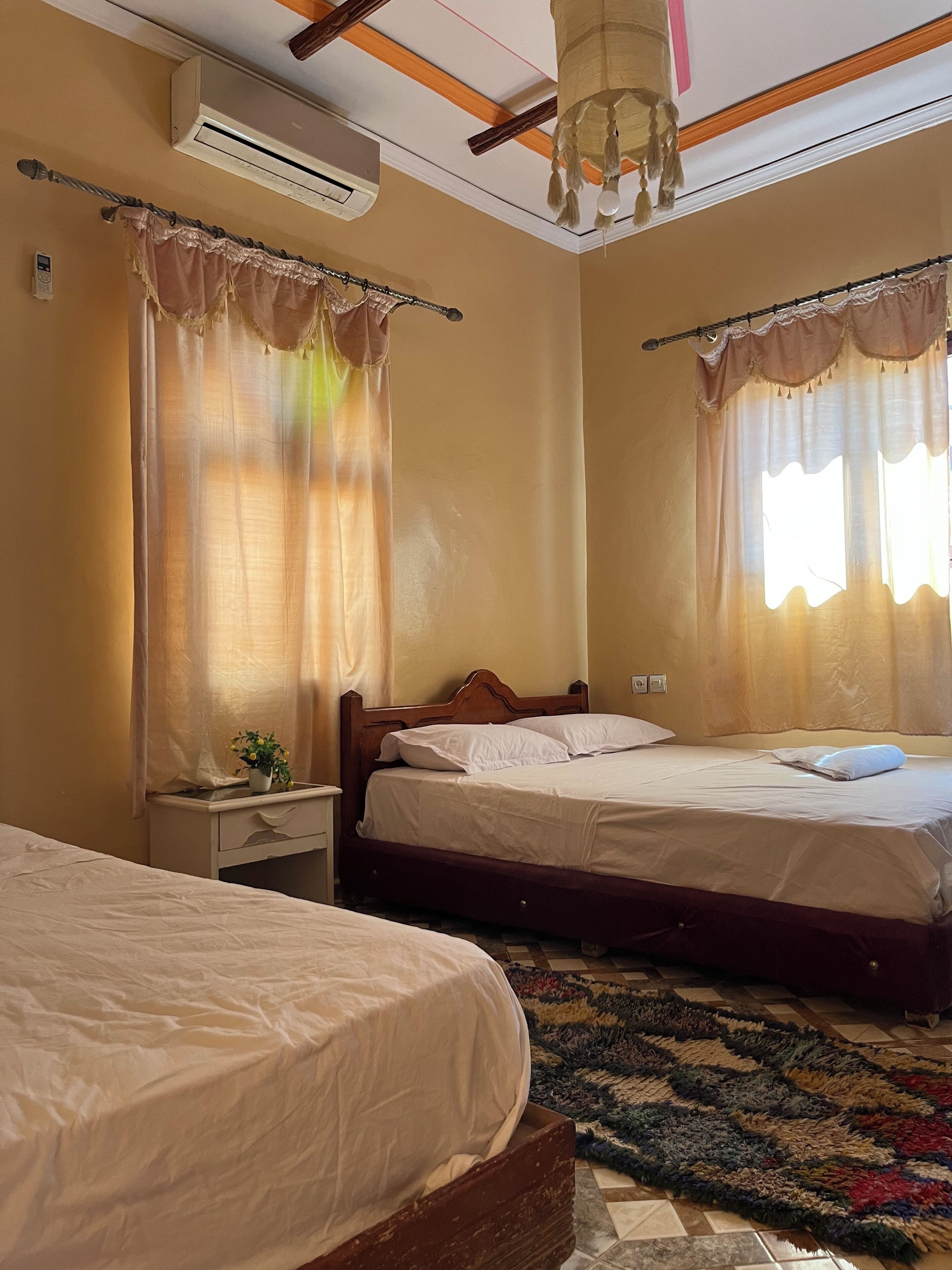 Een hotelkamer met twee bedden, een klein nachtkastje met een lamp, een airconditioner en een plafondventilator.