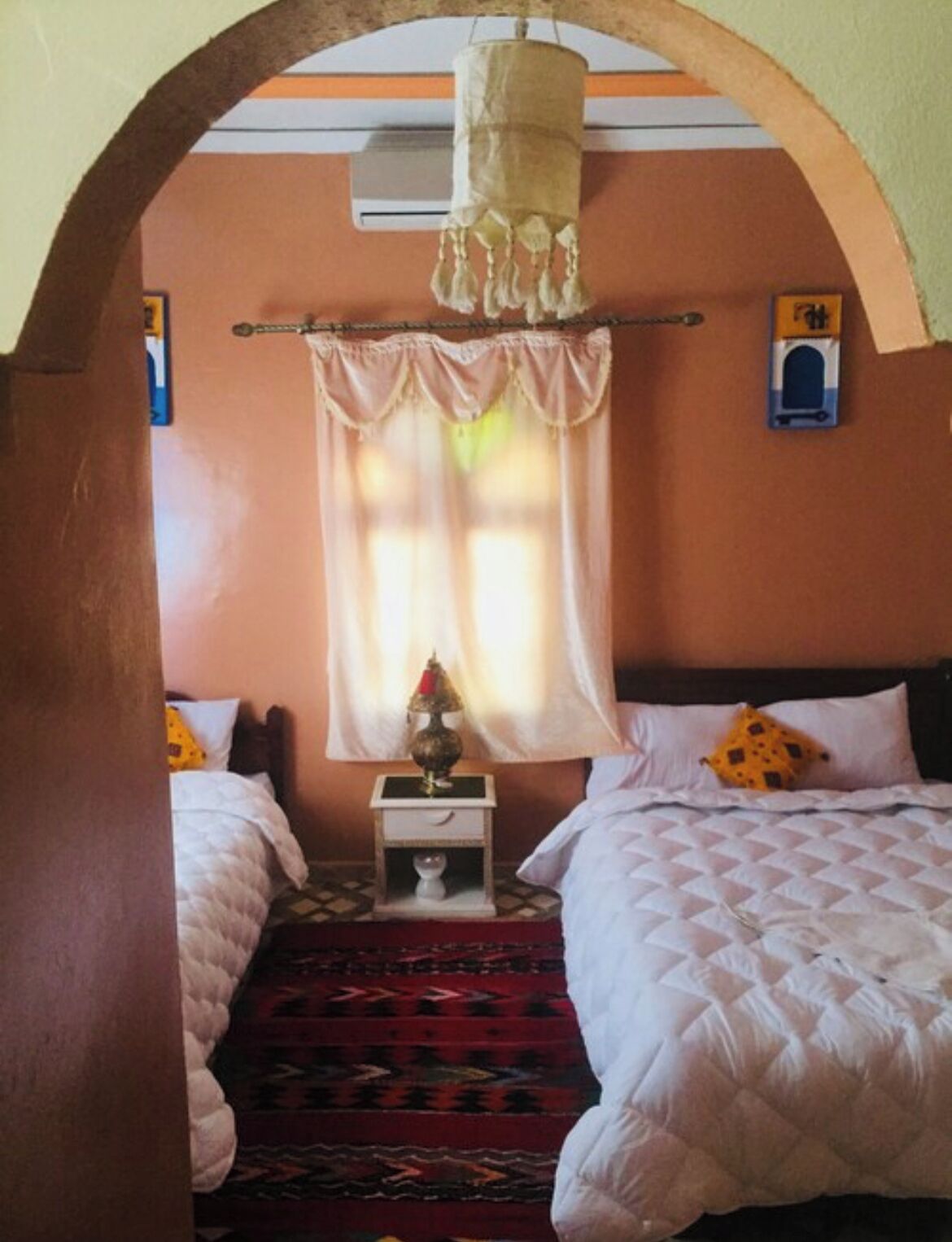 Ein Zimmer mit zwei Betten, einem Nachttisch mit Lampe und einem Fenster mit Vorhängen.