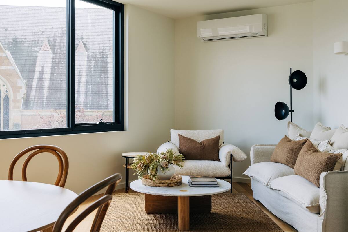 Un salon moderne avec une grande fenêtre, un canapé blanc, une table basse ronde et un climatiseur fixé au mur.