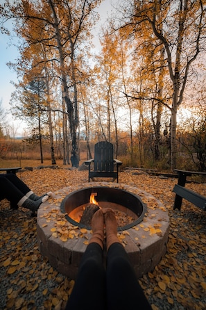 A circular fire pit with a person sitting by it, surrounded by fallen leaves, with trees in the background.