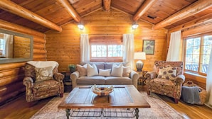 A cozy log cabin interior with a wooden ceiling, a central table, two patterned armchairs, a sofa, and a window with a view of trees. - 20 min to Yellowstone, Family Cabin, Fire Pit (Island Park)