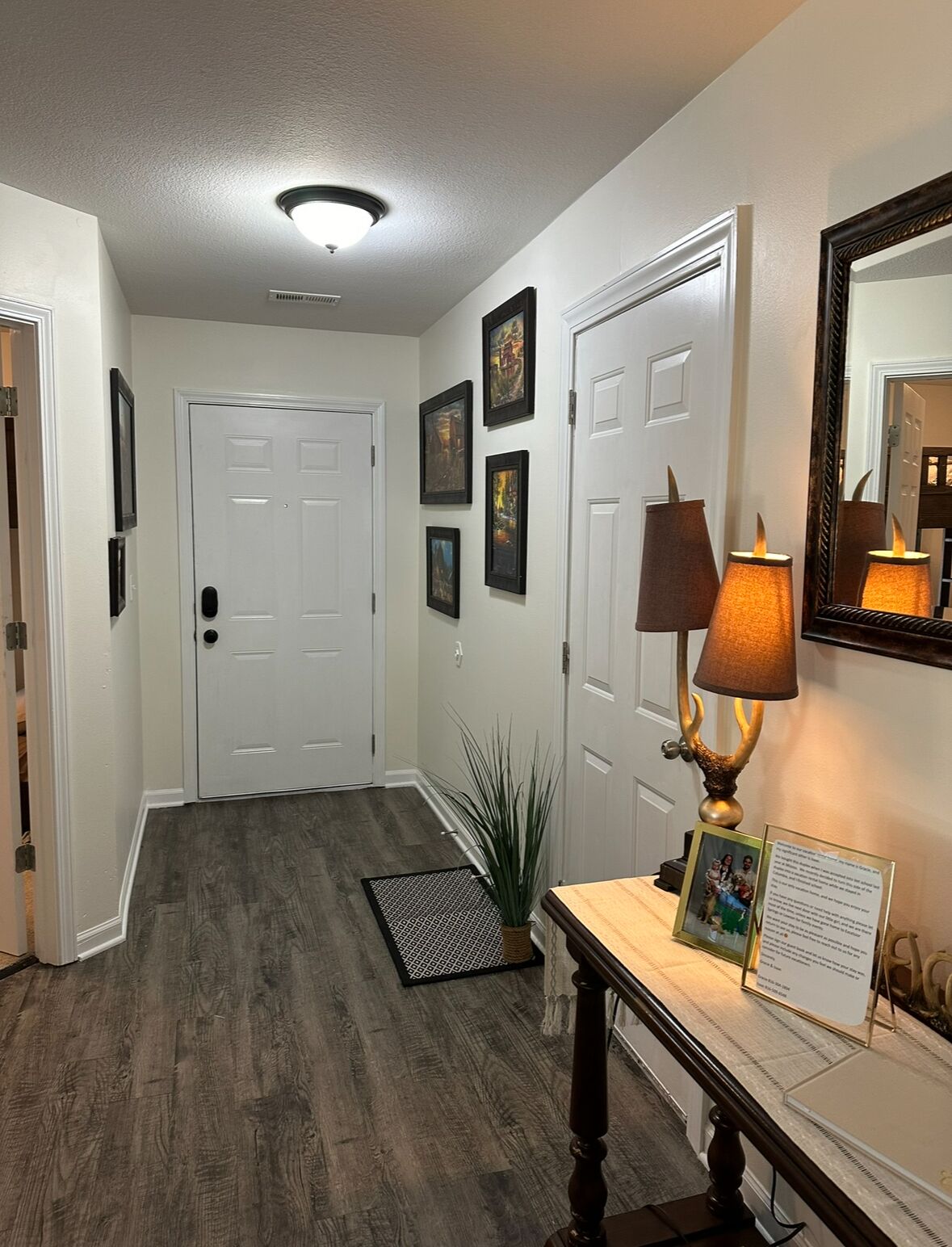 A hallway with a wooden floor, white walls, and a ceiling light. There are framed pictures on the wall, a door, and a small table with a lamp and a framed photo.