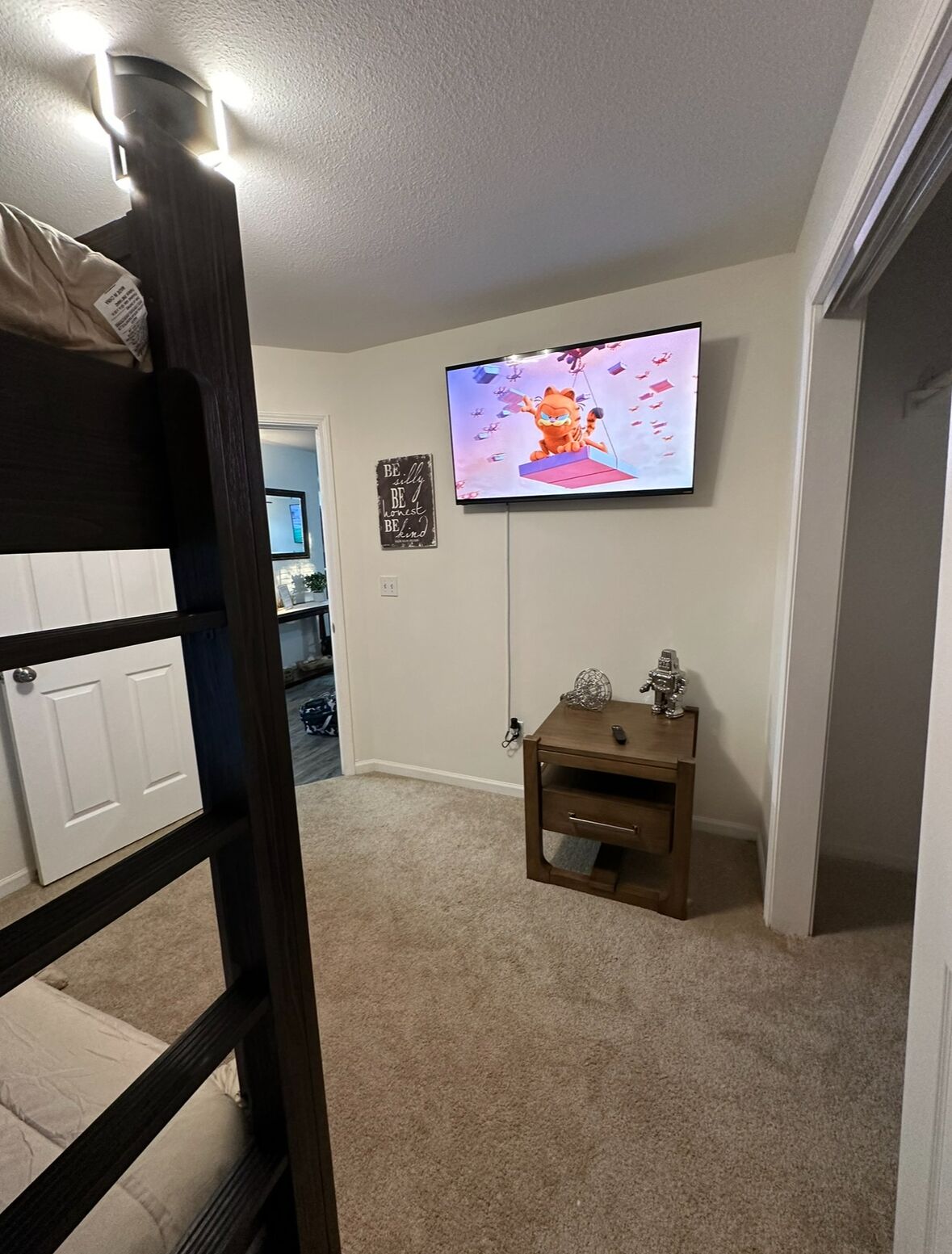 A room with a bunk bed, a television mounted on the wall, a small wooden nightstand, and a motivational quote sign.