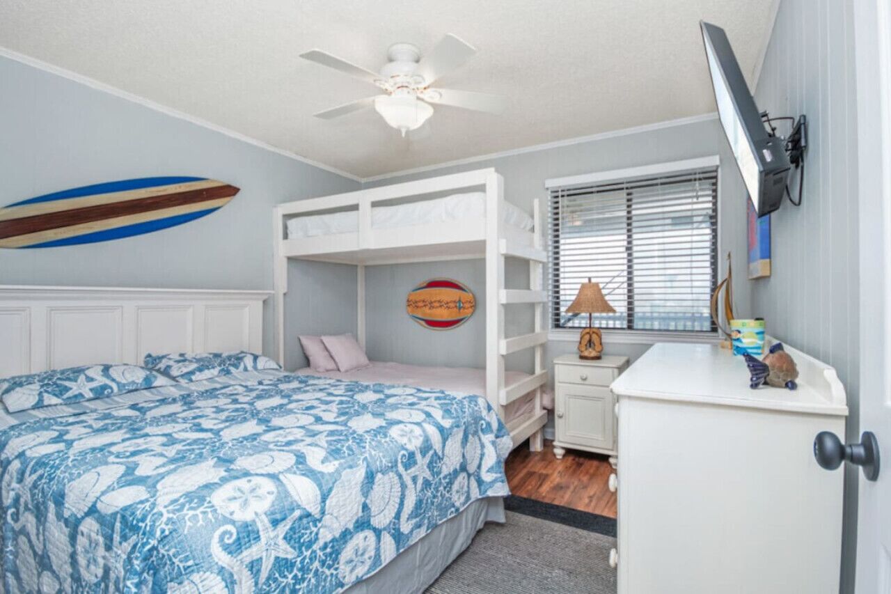 Une chambre avec un lit superposé, un ventilateur de plafond, une décoration sur le thème du surf, un tableau rond accroché au mur, une lampe et un téléviseur posé sur un meuble.