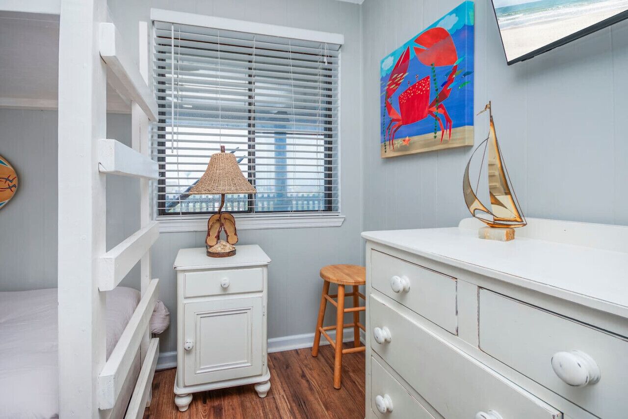A room with a bunk bed, a dresser, a lamp, a wooden stool, and a painting of a crab.