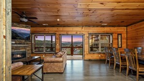 A log cabin interior with a fireplace, seating area, dining table, and large windows offering scenic views.