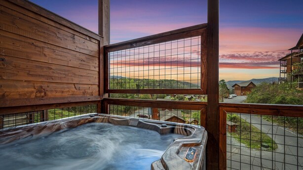 A wooden cabin with a hot tub on a balcony overlooking a scenic landscape during sunset.