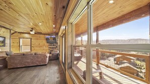 A cozy cabin interior with a wooden ceiling, a fireplace, a sofa, and a balcony with a view of the surrounding landscape.