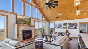 A spacious living room with a fireplace, large windows, and a high ceiling fan.