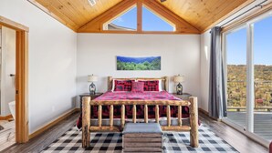 A bedroom with a wooden bed frame, checkered rugs, a painting of a landscape, and a view of a forested area through a large window.