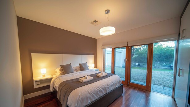 A modern bedroom with a large bed, bedside lamps, and a view through glass doors to an outdoor area. - The Hideaway by Gatt Wines (Flaxman Valley)