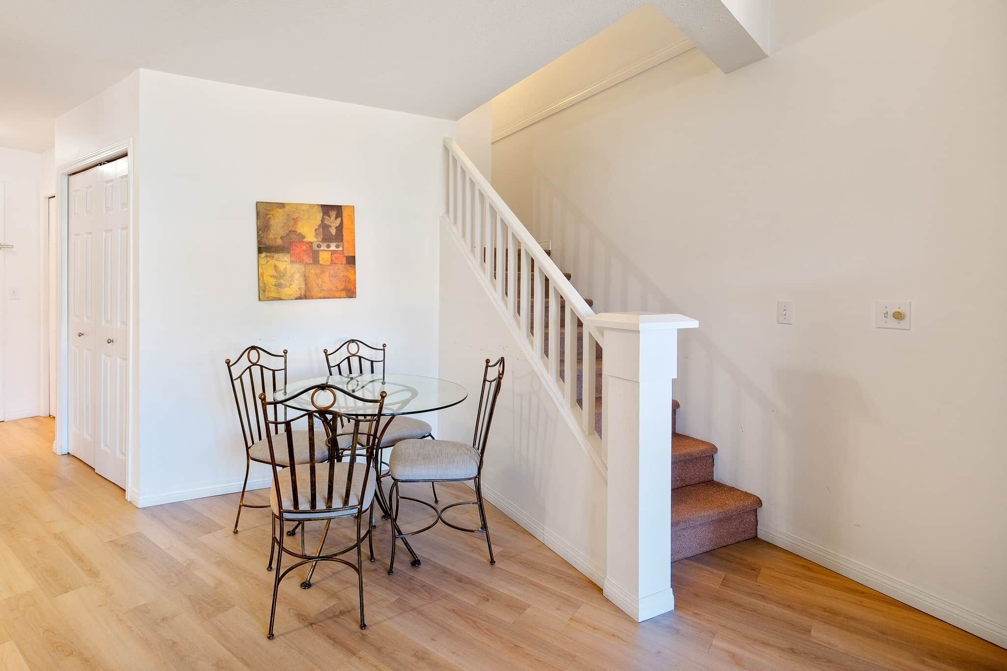 A dining area with a glass table and metal chairs, a staircase with white railings, and a door leading to another room.