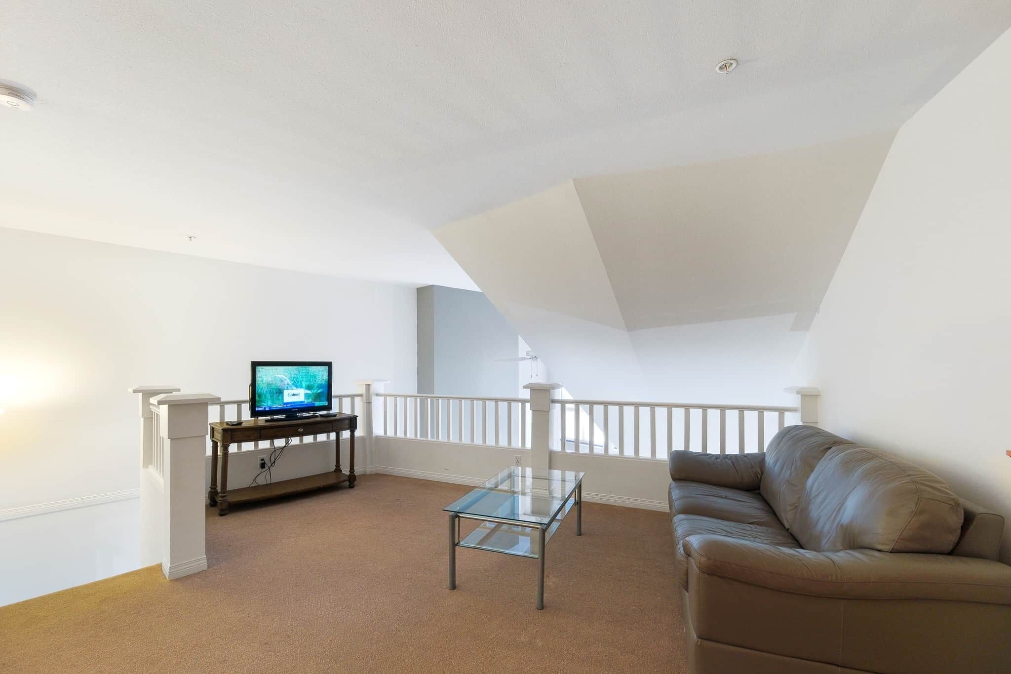 A living room with a glass coffee table, a brown leather sofa, a television on a stand, and a white railing.