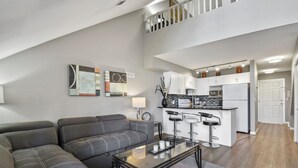 Un salon moderne avec un canapé d’angle gris, une table basse en verre et un coin cuisine doté de meubles blancs et d’un tabouret de bar.