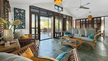 A spacious living area with a large sofa, a coffee table, and wicker furniture. There is a ceiling fan and a decorative hanging light.