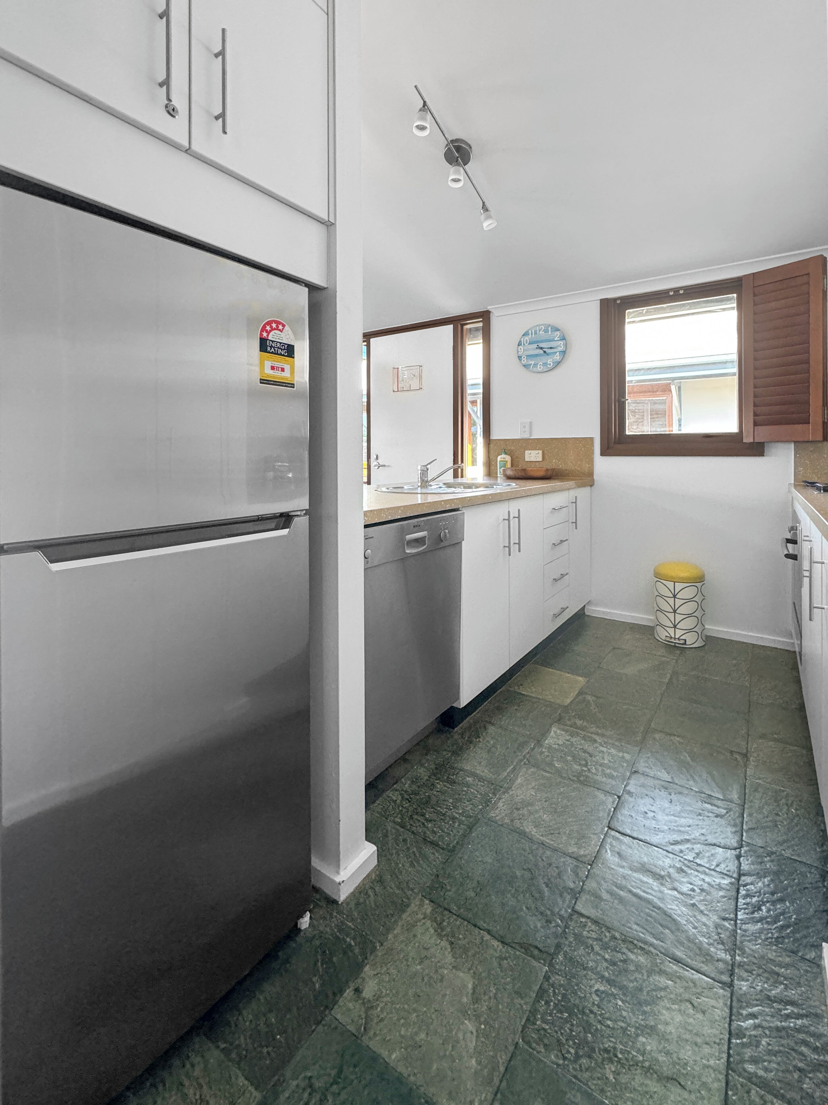 A kitchen with white cabinets, a stainless steel refrigerator, a clock, and a window with wooden shutters.
