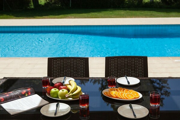A dining table with plates of fruit and drinks, two chairs, and a view of a swimming pool. - Luxurious Villa Retreat with Panoramic Views (Paphos)