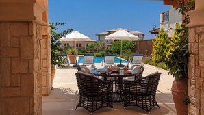 A patio area with a round table and four chairs, a pool, and a building with a tiled roof in the background. - Luxury Villa Retreat with Unmatched Elegance and Serenity (Paphos)