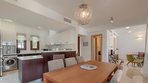 A modern kitchen with white cabinets, a central island, and a dining area with a wooden table and chairs. - Luxury Villa Retreat with Unmatched Elegance and Serenity (Paphos)