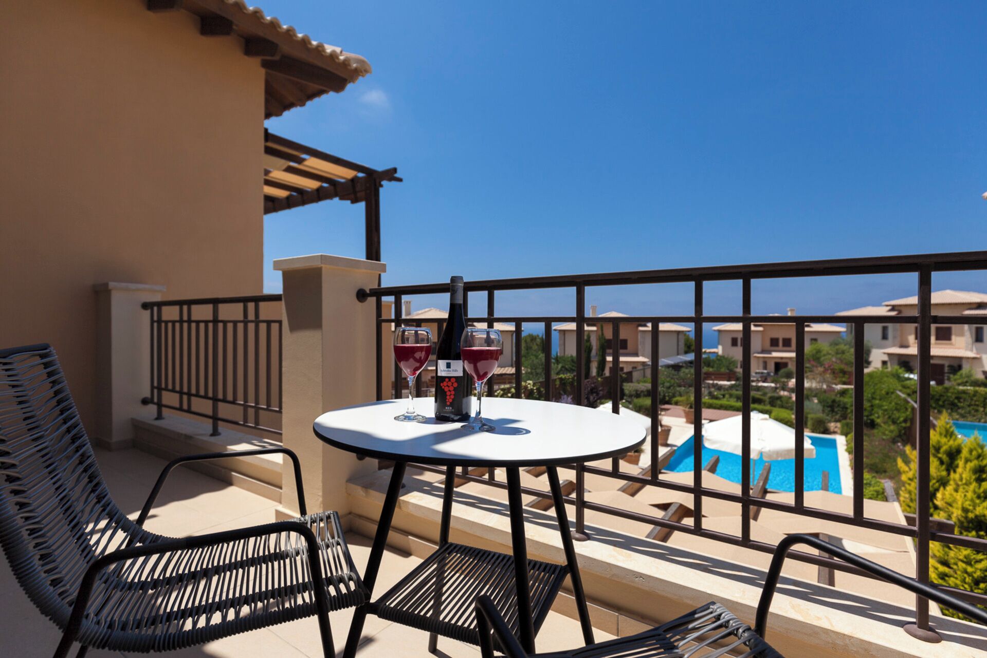 Ein Balkon mit Tisch, Stühlen und einer Weinflasche. Im Hintergrund ist ein Pool zu sehen.